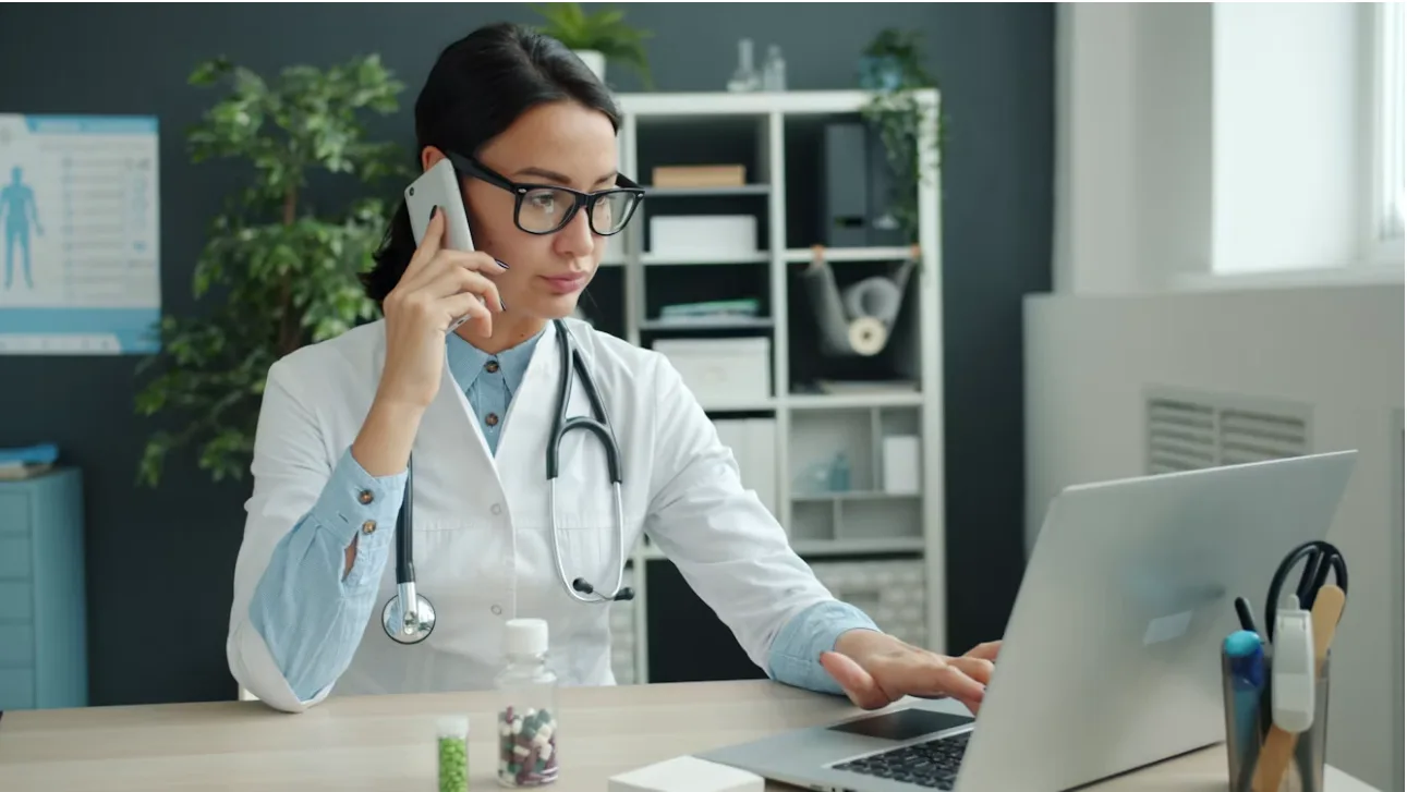 A healthcare professional talking on the phone while working at a laptop.